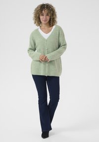 Cardigan tricoté vert clair avec un col en V, des boutons sur le devant et des manches longues, associé à un jean bleu foncé évasé et des bottines noires.