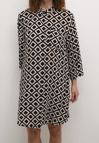 Kortærmet kjole i sort og beige med diamantmønster, med knaplukning foran og krave samt en brystlomme på venstre side.