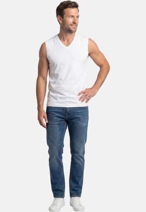 Man staat met de hand op de heup, draagt een witte mouwloze V-hals shirt, blauwe spijkerbroek en witte sneakers, tegen een effen achtergrond.