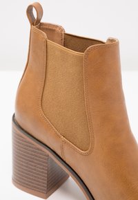 Botín de cuero color tostado con paneles laterales elásticos y un tacón cuadrado marrón texturizado. Cuenta con una pestaña de agarre para facilitar su uso.