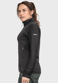 Schwarze Zip-Jacke mit einem hohen Kragen und gerippter Textur. Verfügt über ein Logo am Ärmel und eine mit Reißverschluss ausgestattete Seitentasche.