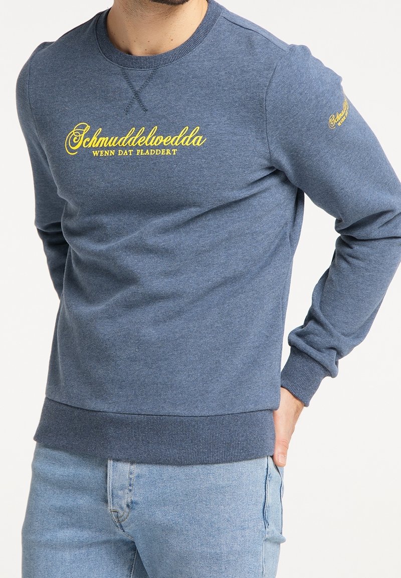 Sweat-shirt bleu à manches longues avec texte jaune « Schmuddelwedda Wenn Dat Pladdert » porté par une personne avec une main dans la poche d’un jean bleu clair.