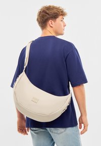 Béžová crossbody taška z textilu, s zakriveným dizajnom, nastaviteľným popruhom a malým logom. Nosená cez rameno.