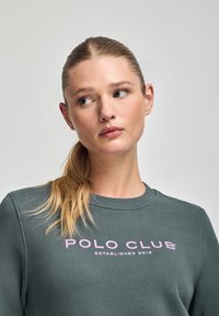 Sudadera gris, con cuello acanalado, que presenta el texto en rosa "POLO CLUB" y "ESTABLECIDO 2012" abajo. Textura suave, diseño casual.