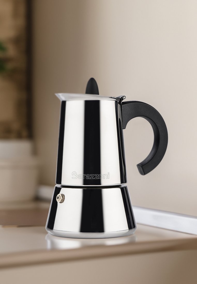 Cafetière moka en acier inoxydable avec une poignée en plastique noir. Elle présente une base ronde, un corps conique et une finition métallique avec un logo à l'avant.