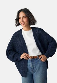 Cardigan en maille épaisse bleu marine, design à ouverture avant avec manches larges. Associé à une chemise blanche et un jean en denim taille haute, tissu texturé.