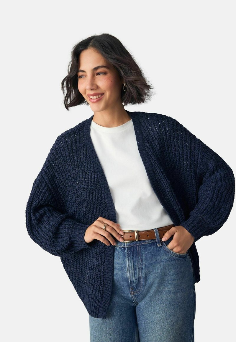 Cardigan en maille épaisse bleu marine, design à ouverture avant avec manches larges. Associé à une chemise blanche et un jean en denim taille haute, tissu texturé.