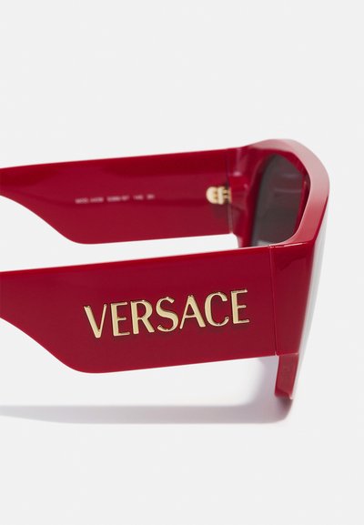 Ochelari de soare roșii din plastic au un design voluminos, cu lentile întunecate și un logo „VERSACE” proeminent pe lateral.