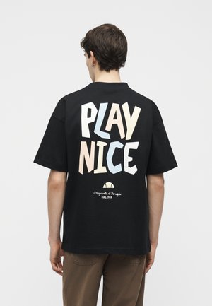 Νέος άνδρας με κοντά μαλλιά που φοράει μαύρο φαρδύ T-shirt με την επιγραφή "PLAY NICE" σε παστέλ χρώματα στην πλάτη, συνδυασμένο με καφέ παντελόνι.