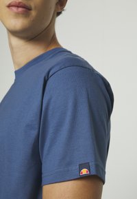 Niebieska bawełniana koszulka z okrągłym dekoltem i krótkimi rękawami. Na lewym rękawie znajduje się małe, wielokolorowe logo. Materiał o średniej grubości.