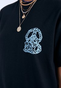 Camiseta de algodón negra con un gráfico de símbolo de paz floral en azul claro en el pecho. Textura suave, mangas cortas y cuello redondo.