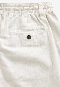 Pantalons en lin beige avec taille élastique, passants de ceinture et une poche arrière fermée par un bouton noir.