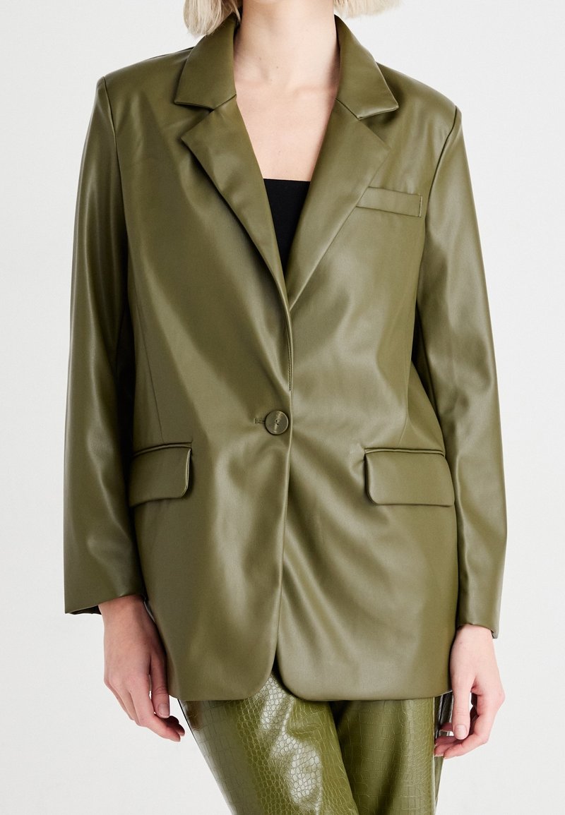 Femme portant un blazer en simili cuir vert olive avec des revers crantés, un bouton devant, des poches à rabat et un pantalon texturé assorti.