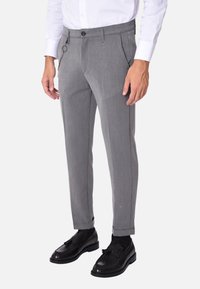 Pantaloni grigi su misura con una texture liscia, dotati di zip, tasche laterali e un anello per una catena, abbinati a mocassini neri.