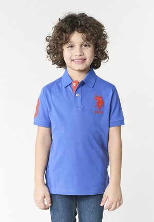 Bambino sorridente con capelli ricci che indossa una polo blu USPA e jeans, in piedi contro uno sfondo bianco semplice.