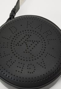 Borsa a tracolla rotonda nera in pelle con logo del designer perforato e tracolla regolabile su uno sfondo bianco.