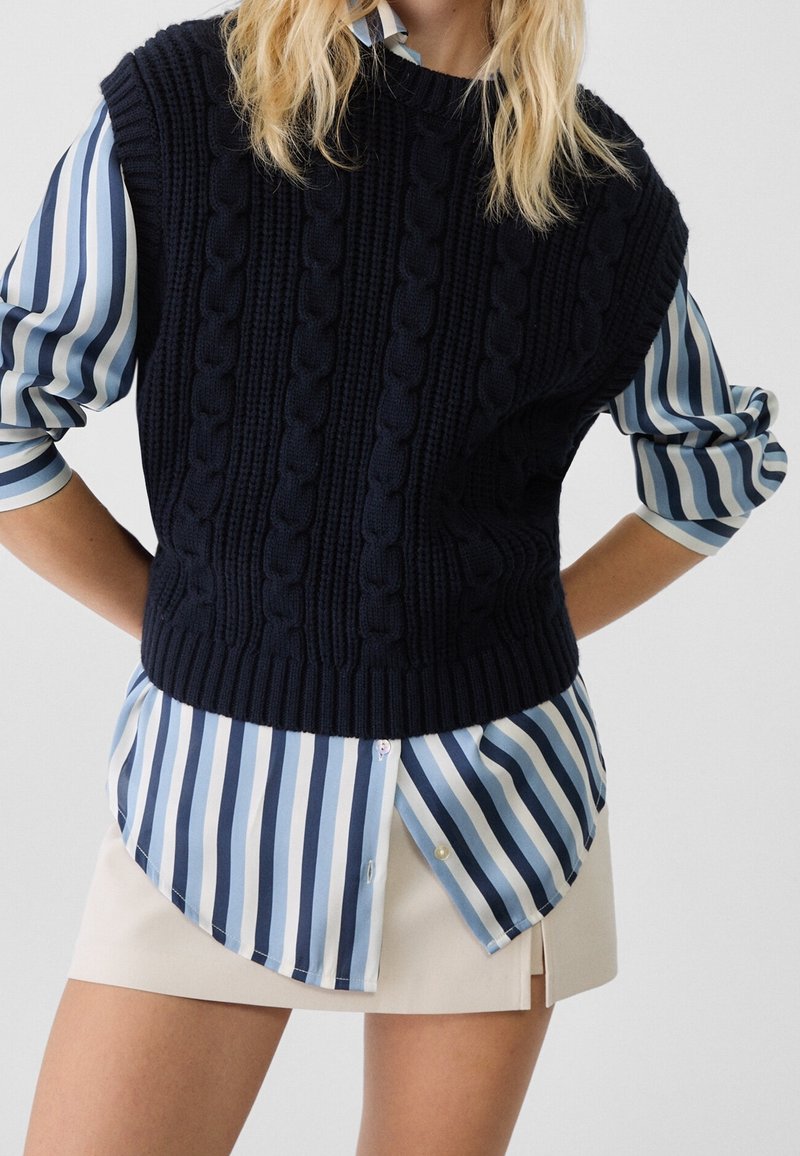 Blond person i en marinblå kabelstickad ärmlös tröja över en blå och vit vertikalt randig skjorta och en beige minikjol.