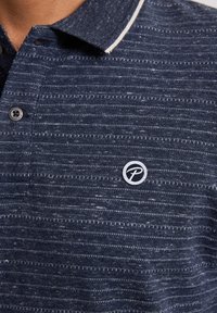 Camisa polo azul marino con un patrón rayado texturizado, que presenta un cierre de dos botones y un logo circular en el pecho. Acentos en el cuello en blanco.
