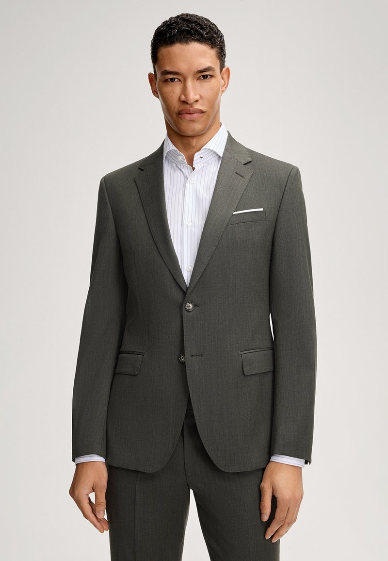 Mężczyzna w ciemnoszarym garniturze na dwa guziki z białą koszulą w paski i białą poszetką, stojący na jednolitym tle.