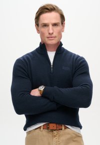 Maglione blu marina con design a mezza zip, polsini a costine e un logo sottile. Indossato sopra una camicia bianca, abbinato a pantaloni kaki e una cintura marrone.