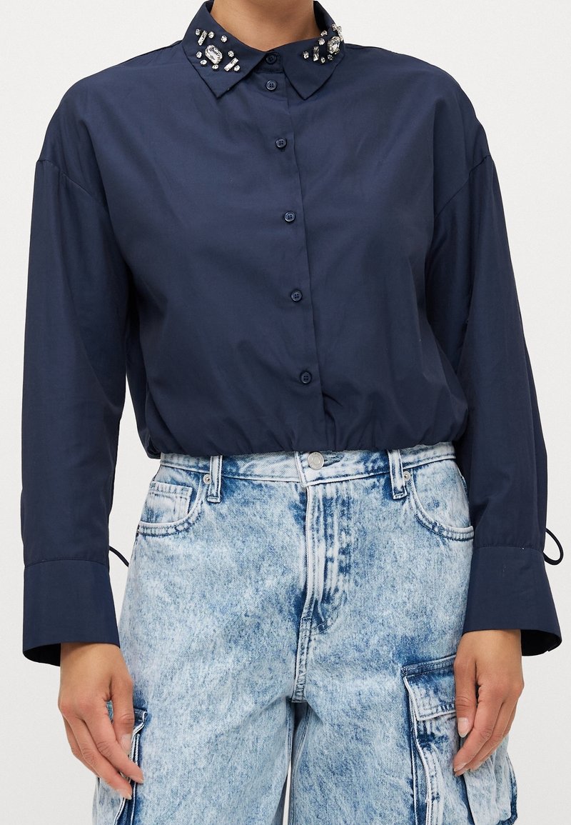 Chemise bleu marine à boutons avec col orné, associée à un jean cargo taille haute délavé clair.