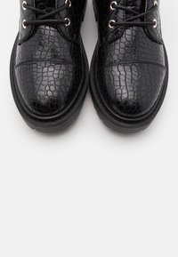 Bottines noires à lacets en matériau brillant et texturé avec un motif de cuir de crocodile. Présente un orteil arrondi et une semelle noire robuste.