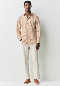 Camisa de botones floral en color crema con patrones rojos y verdes, combinada con pantalones beige claros y zapatos marrones tipo mocasín. Diseño minimalista.