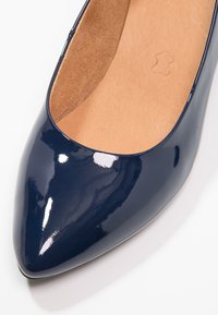 Mocassin en cuir verni bleu marine avec un bout pointu et un extérieur lisse. L'intérieur est doublé en suede tan. Aucun matériel ou motif visible.