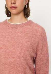 Svetlo roza pletena jopica s okroglim izrezom, izdelana iz mehke teksturirane tkanine. Ima raglan rokave in rahlo sproščen kroj.