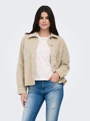 Vrouw met lang donker haar die een beige corduroy jas, wit T-shirt en gescheurde blauwe spijkerbroek draagt, staande met één hand in de zak.