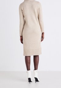 Femme vue de dos portant une robe midi à manches longues beige et des bottines à talons hauts blanches avec des boucles.