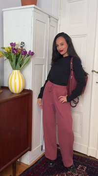 Γυναίκα με μαύρο μακρυμάνικο τοπ και βουργουνδί παντελόνια στέκεται μέσα σε εσωτερικό χώρο κοντά σε ένα λευκό ντουλάπι με βάζο λουλουδιών με κίτρινες ρίγες.