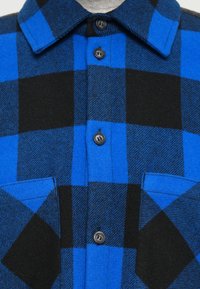 Camicia a quadri blu e nera realizzata in tessuto morbido. Presenta un colletto a punta, chiusura con bottoni e due tasche sul petto. Motivo testurizzato su tutta la superficie.