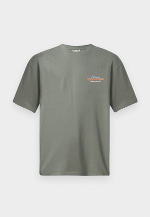 Olīvu zaļš kokvilnas T-krekls ar īsām piedurknēm un apaļu kakla izgriezumu. Uz kreisās krūts zonas ir neliela krāsaina teksta druka.