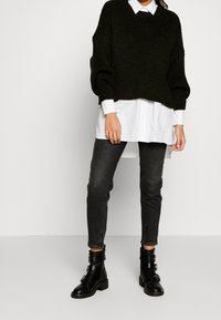 Maglione lavorato a maglia verde scuro sovrapposto a una camicia bianca con colletto, abbinato a jeans grigi scuri e stivaletti neri alla caviglia con fibbie.