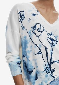 Pull en maille avec col en V, base blanche et imprimé floral bleu. Manches longues avec poignets texturés et tissu doux et léger.