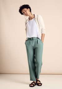 Helles beiges Cardigan über einem weißen Oberteil, kombiniert mit mintgrünen, hochtaillierten Hosen und schwarzen Sandalen mit gemusterten Riemen.