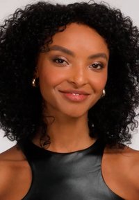 Donna con capelli ricci neri che indossa orecchini a cerchio dorati e un top nero senza maniche, che sorride dolcemente contro uno sfondo chiaro.