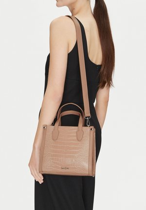 Kobieta w czarnej sukience niosąca beżową teksturowaną skórzaną torebkę z uchwytami na górze i paskiem na ramię przewieszonym przez ramię.