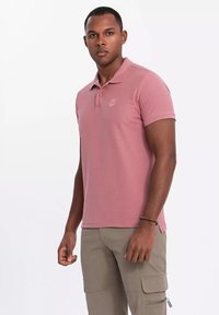 Polo tričko s krátkým rukávem v svetle růžové látce, se límečkem a dvěma knoflíky. Discretni logo na hrudi. Nenošeno s béžovými cargo kalhotami.