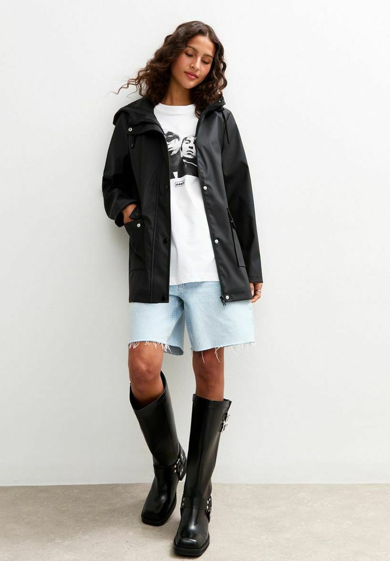 Czarna wodoodporna kurtka z kapturem, biała koszulka z graphic, jasnoniebieskie szorty dżinsowe i wysokie czarne gumowe kalosze z akcentami w postaci klamerek.