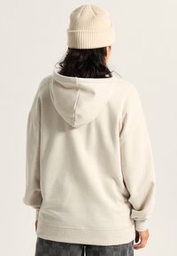 Person trägt einen beigen Hoodie und eine passende Strickmütze, von hinten gesehen, mit dunklen Haaren und grauen karierten Hosen.