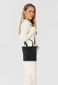 Schwarze Leder-Tote mit zwei kurzen Griffen, minimalistisches Design und einem sichtbar auf der Vorderseite eingeprägten Markenschild.