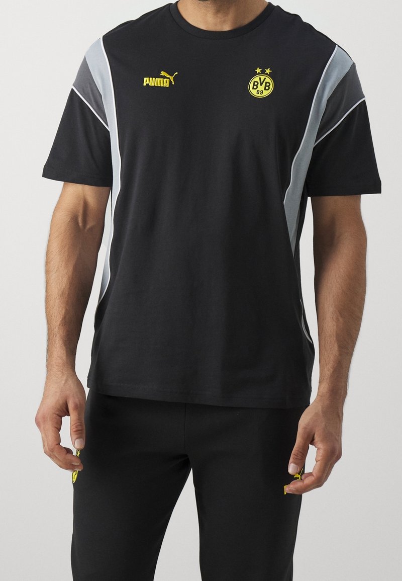 Man som bär svart Puma sportkläder med grå ränder och Borussia Dortmund-logotyper på tröjan och byxorna, stående mot en enfärgad bakgrund.