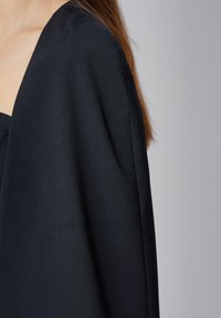 Vêtement noir, à la texture douce, avec un design large et ouvert et un décolleté profond, encadré au niveau de l'épaule, mettant en valeur un tissu lisse et un drapé élégant.