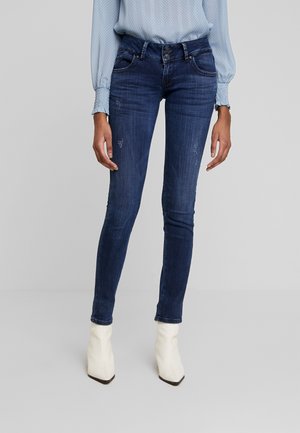 Jeans ajustados en denim azul oscuro, con una cintura de altura media, acentos desgastados y un diseño clásico de cinco bolsillos. Combinados con botas de tacón blancas hasta el tobillo.