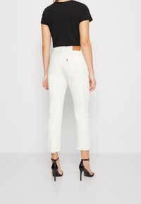Jeans blancos de cintura alta con dobladillos deshilachados, que presentan una etiqueta de cuero en la parte trasera de la cinturilla y un corte recto y ajustado. Top negro y tacones.