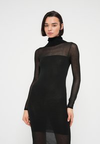 Γυναίκα με κοντά σκούρα μαλλιά που φοράει εφαρμοστό διάφανο μαύρο φόρεμα με ζιβάγκο, στέκεται μπροστά σε λευκό απλό φόντο.