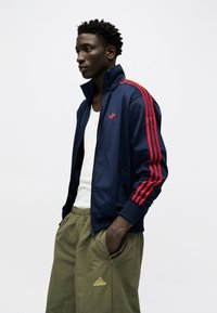 Mann iført en marineblå treningsjakke med røde striper og olivengrønne Adidas-bukser, stående med en hånd i lommen.