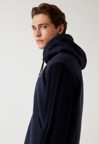 Felpa navy con un design a treccia testurizzato sulle maniche, tasca frontale a marsupio e cappuccio con cordino regolabile. Materiale morbido in cotone.
